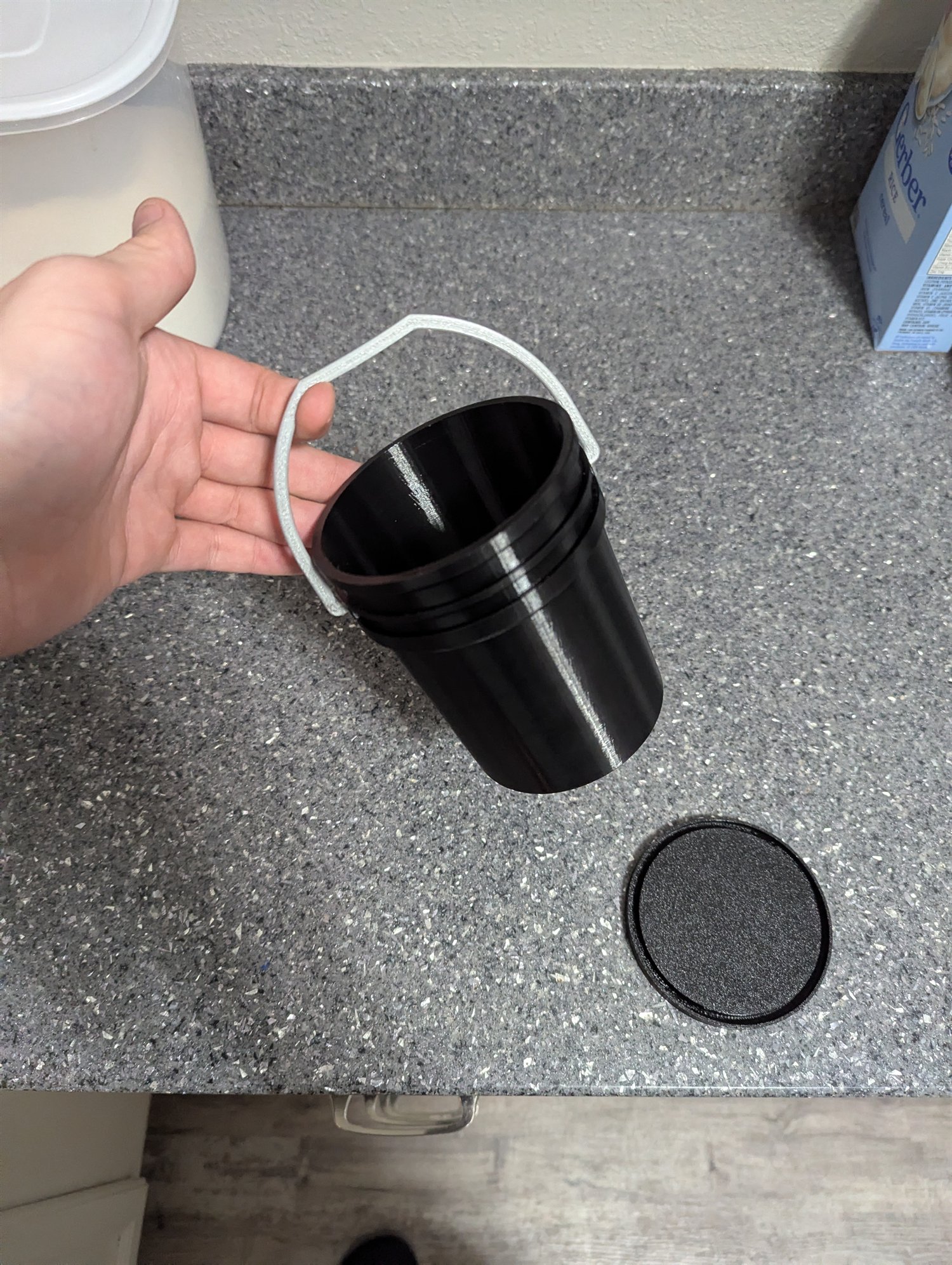 Open black miniature five-gallon bucket held by its gray 3D-printed handle with the lid beside it